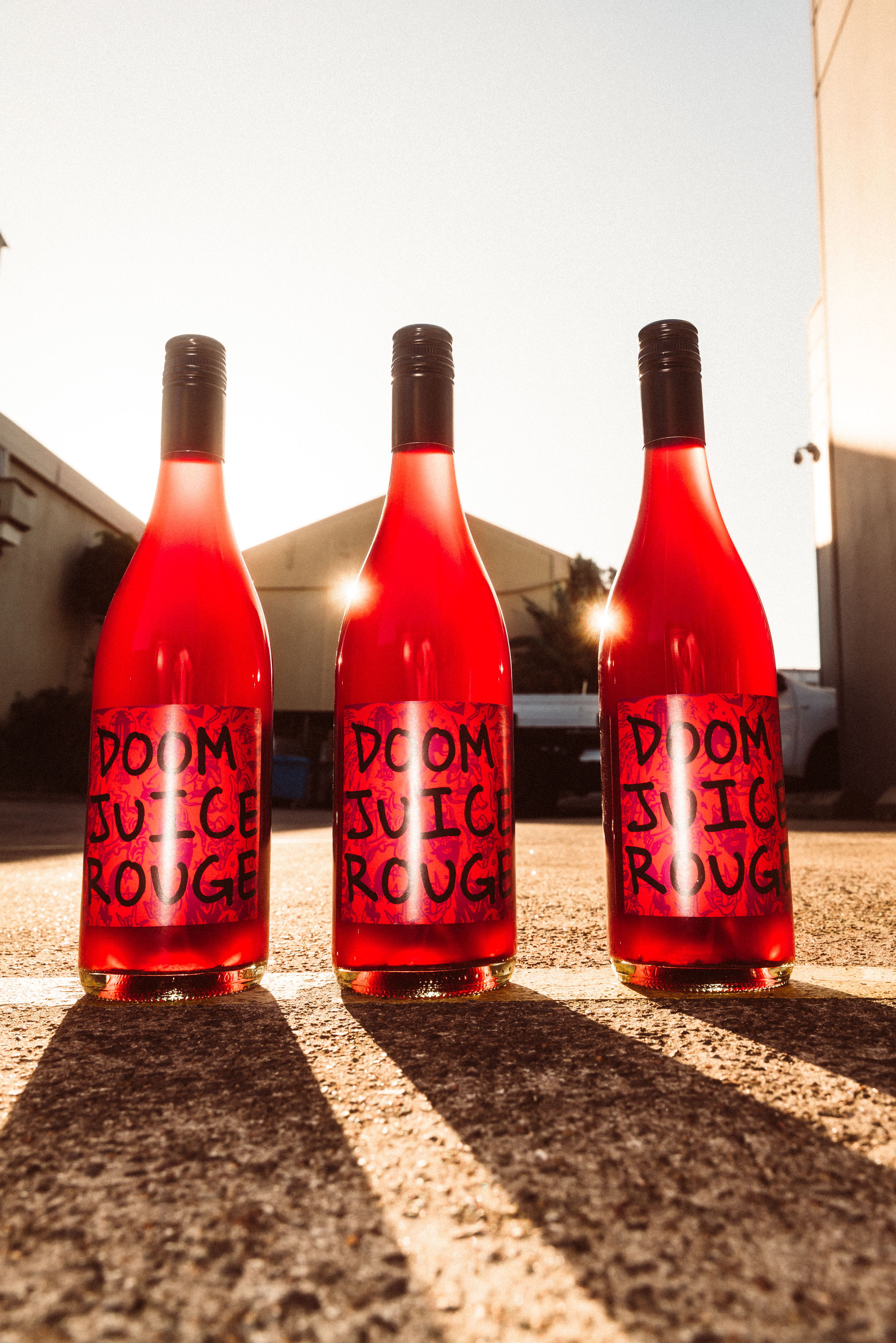 Three bottles of 'Doom Juice Rouge' wine on a concrete surface with a building in the background.