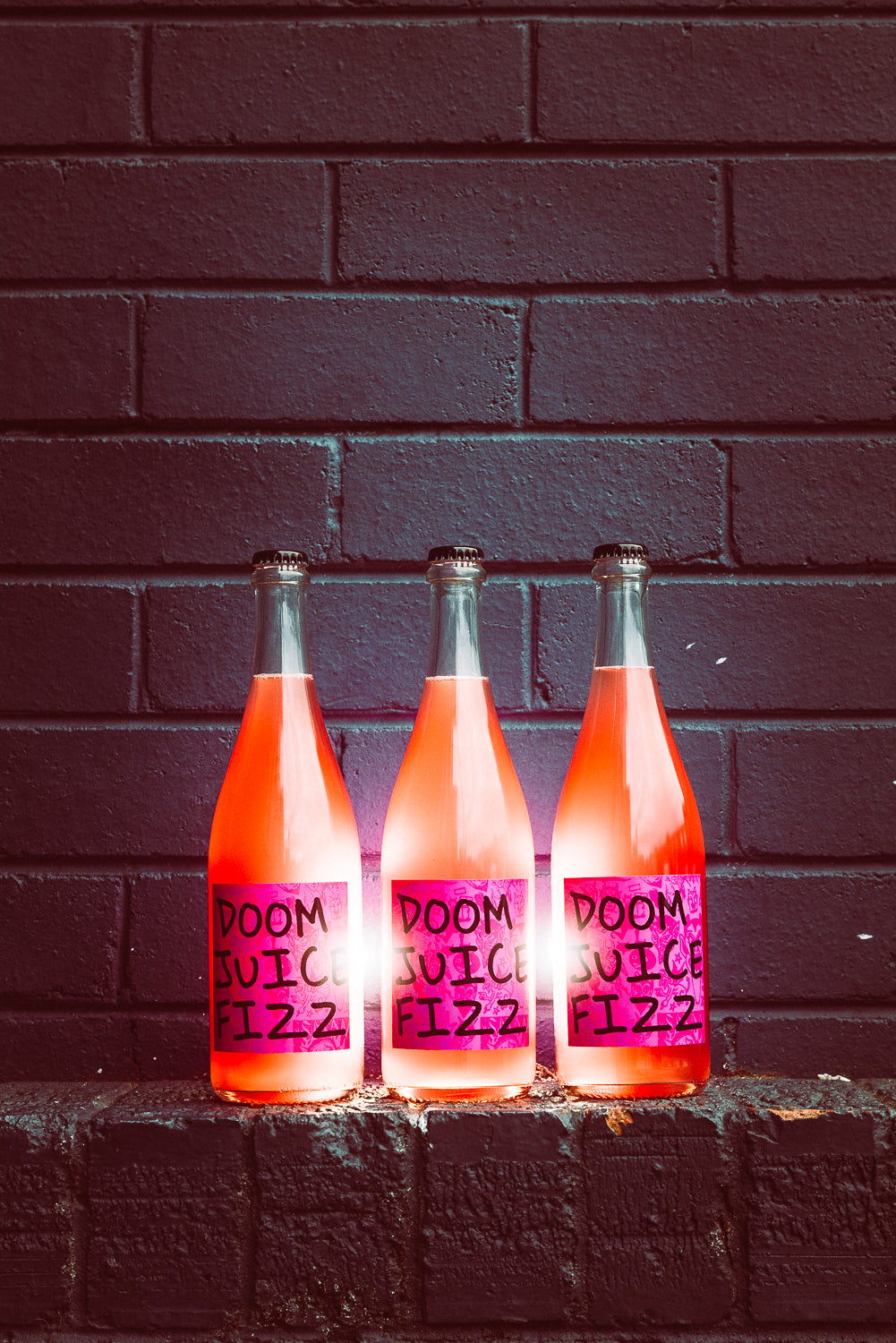Three bottles of 'DOOM JUICE FIZZ' against a dark brick wall.