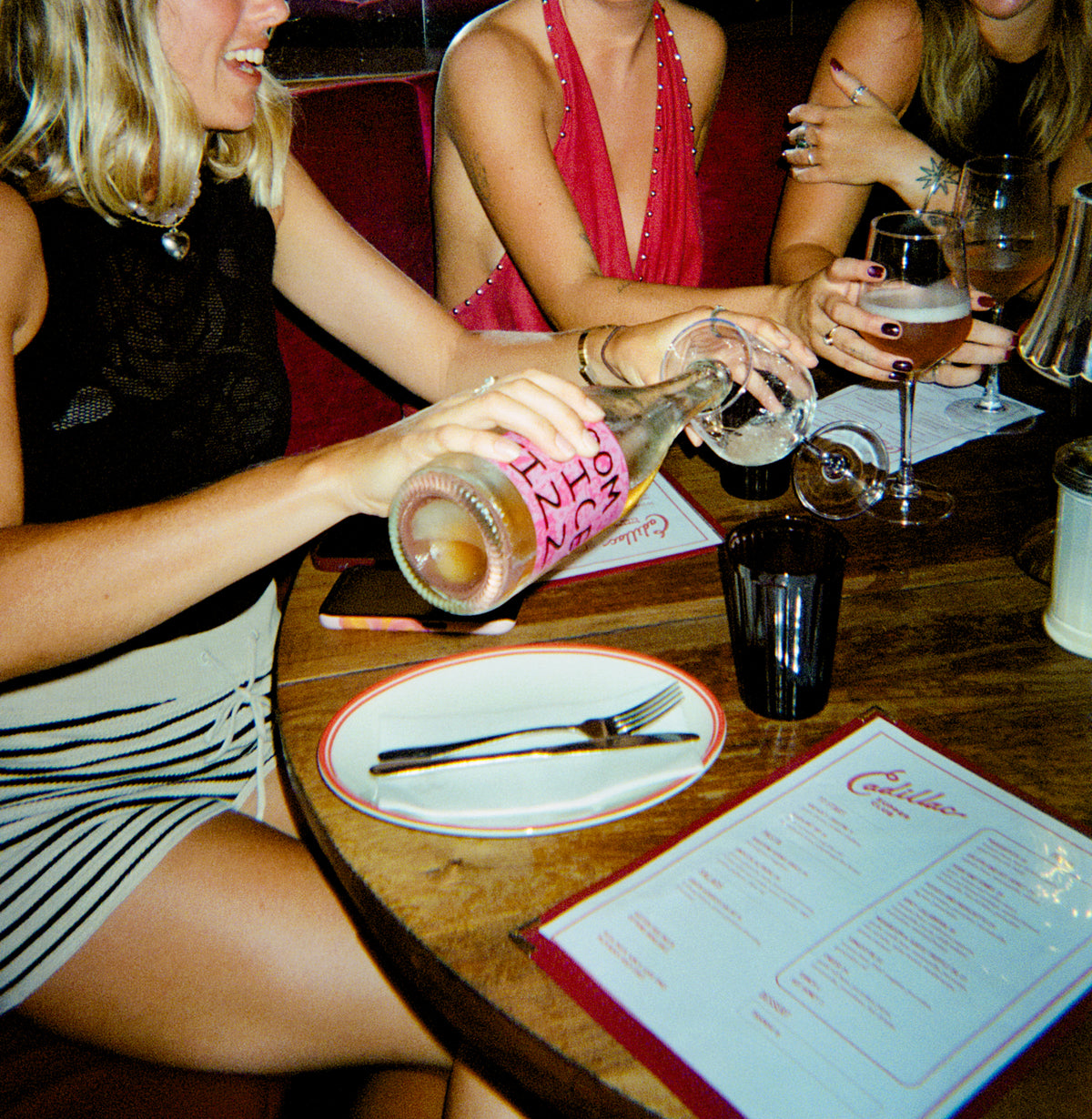 Two women at a bar, one pouring a drink from a bottle labeled 'Doom Juice Fizz' into a glass.