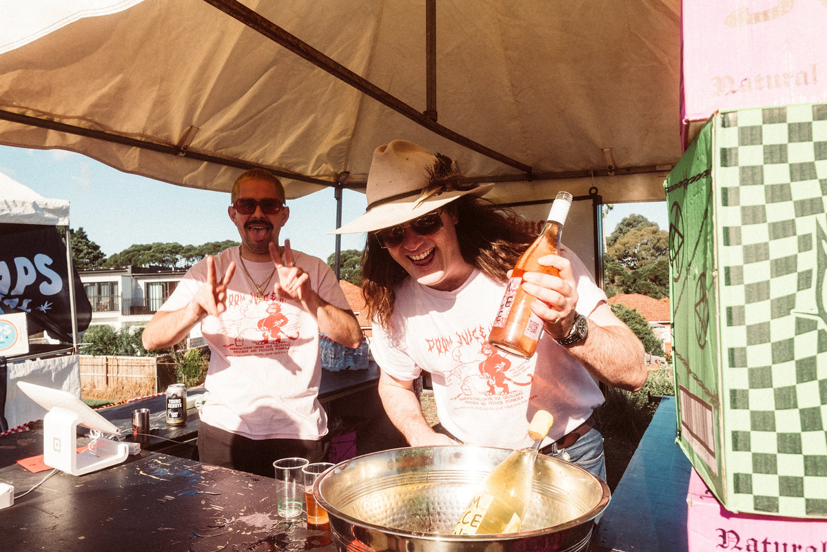 Two people under a large umbrella at an outdoor event, one holding a bottle.